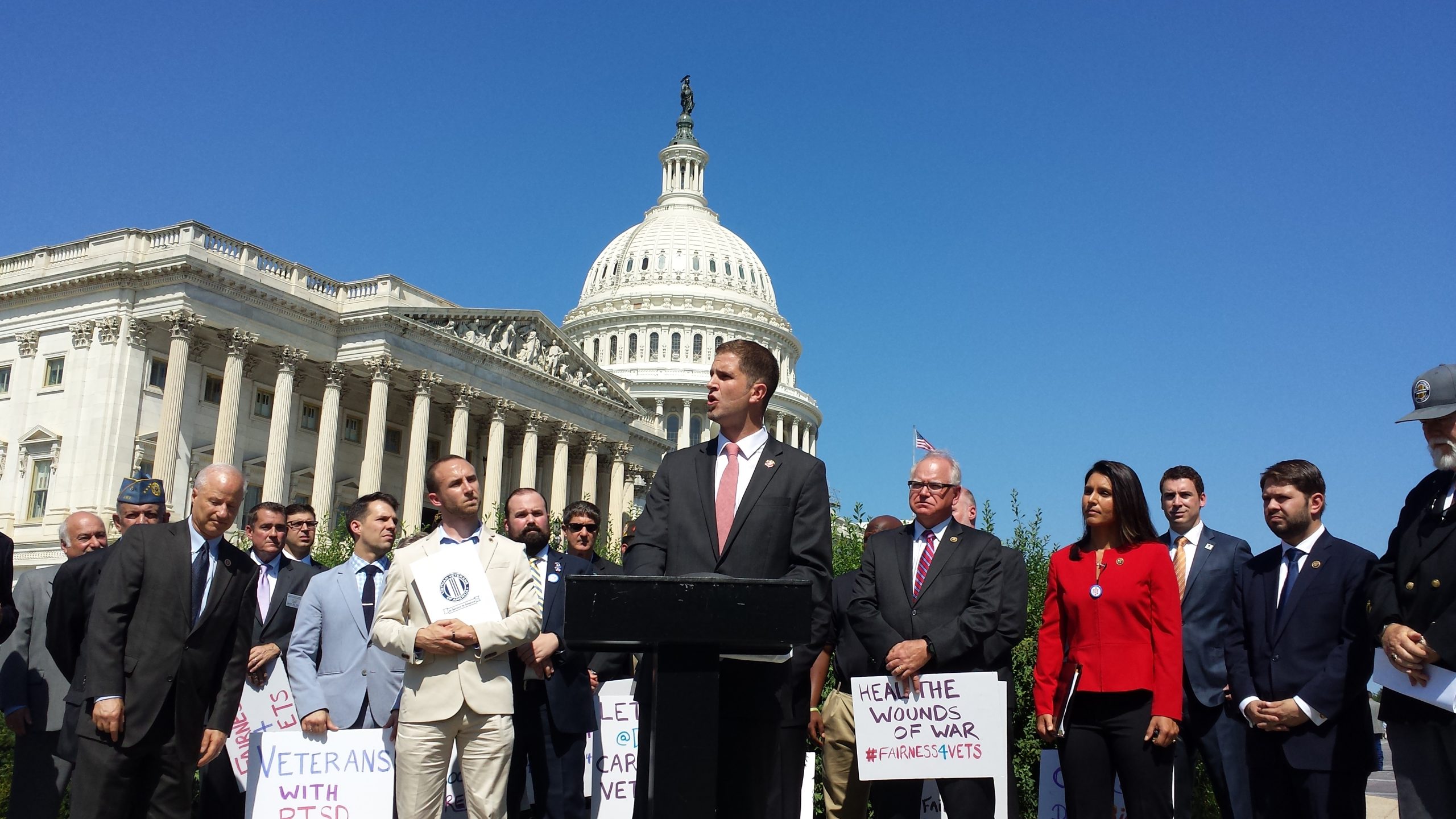 Tyson Manker speaking at a podium in front of the United States Capitol, flanked by members of Congress and veterans holding signs reading Veterans with PTSD and Heal the Wounds of War